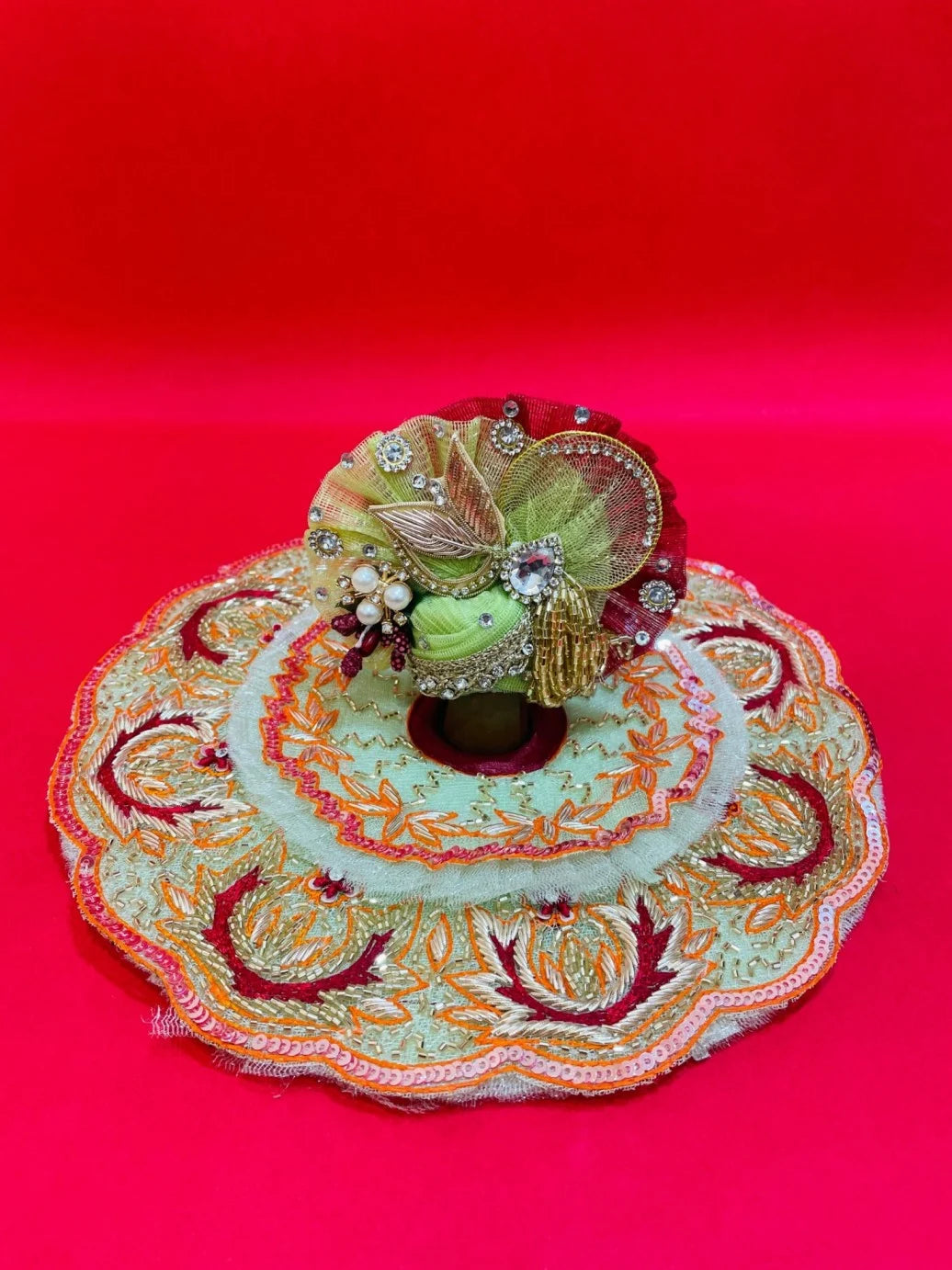 Green and orange embroidered Laddu Gopal dress set with pagdi and patka, displayed on red background and worn by idol.