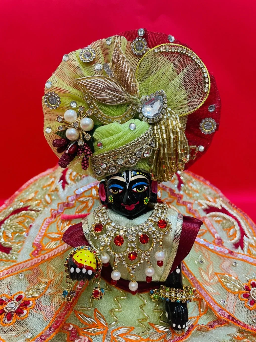 Green and orange embroidered Laddu Gopal dress set with pagdi and patka, displayed on red background and worn by idol.