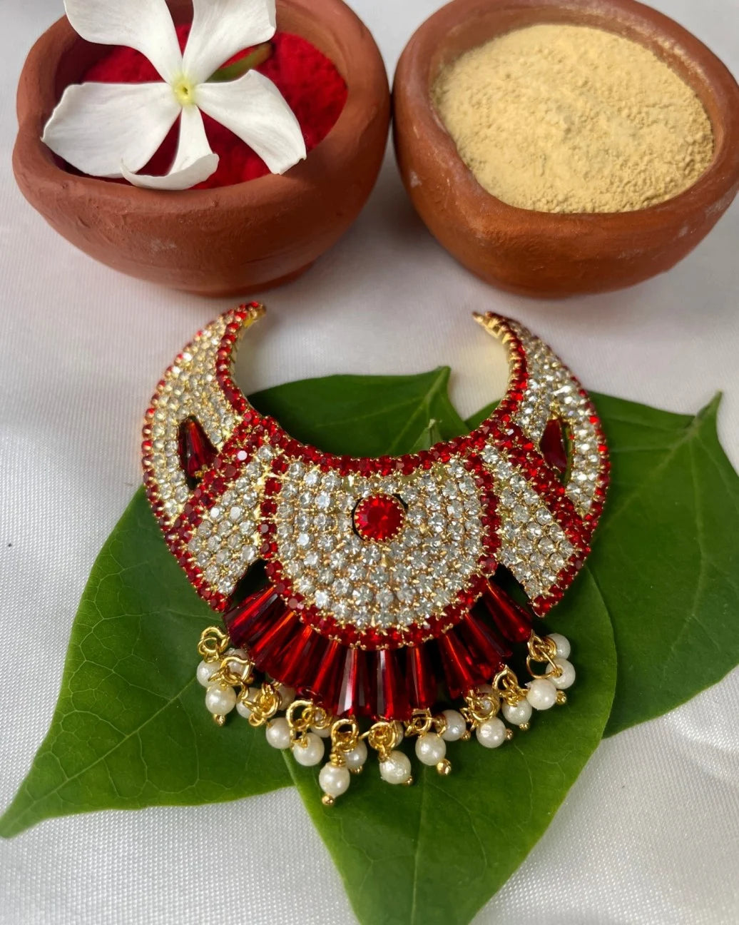 Fan-style red Laddu Gopal jewellery with white stones and pearl hangings placed on green leaves.