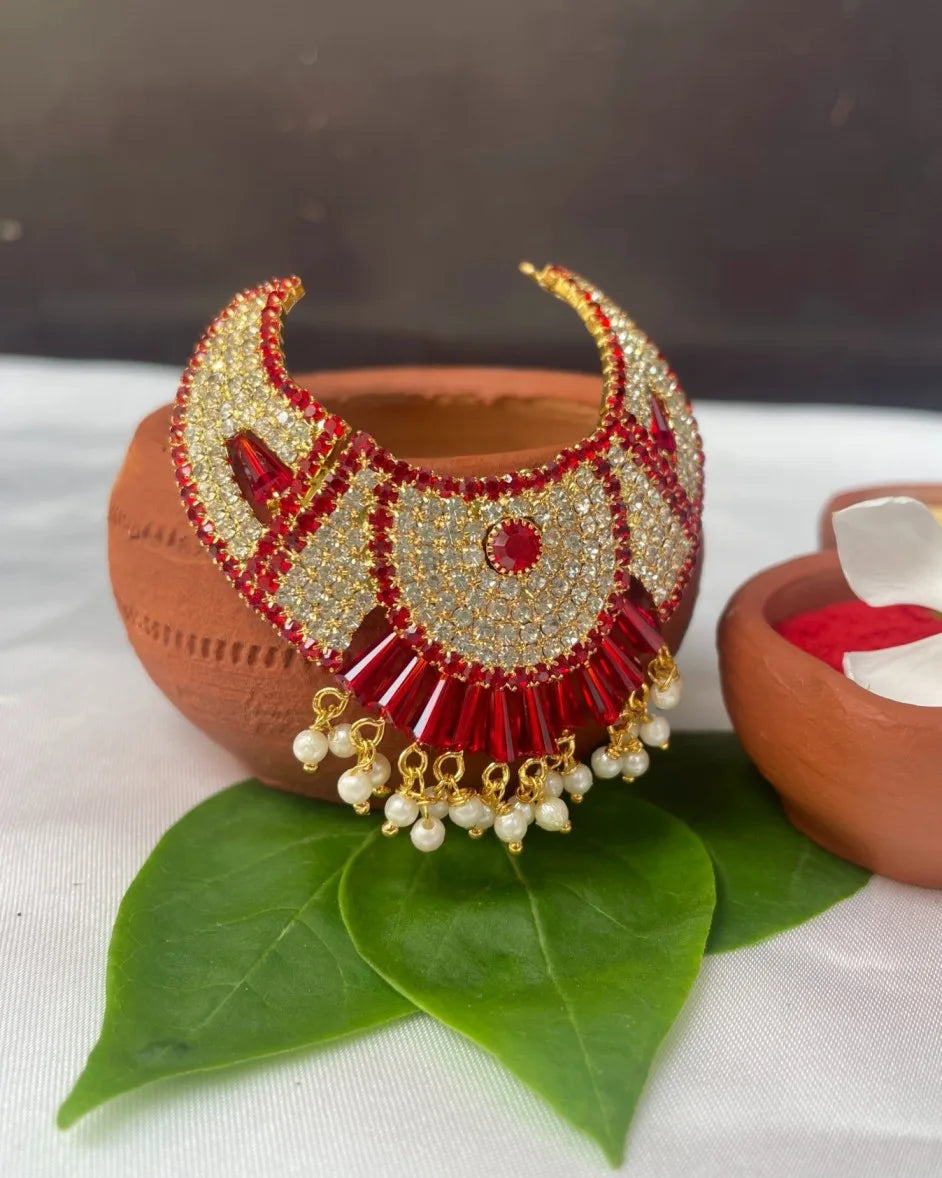 Fan-style red Laddu Gopal jewellery with white stones and pearl hangings placed on green leaves.