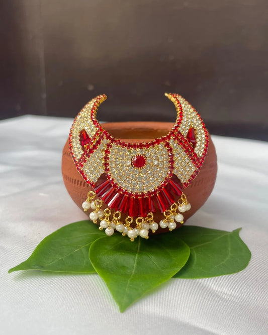 Fan-style red Laddu Gopal jewellery with white stones and pearl hangings placed on green leaves.