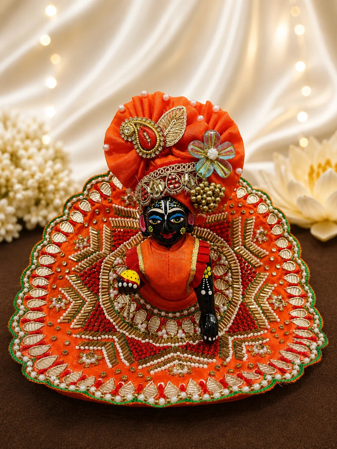 Vibrant orange festive Laddu Gopal poshak set with intricate white pearl beadwork, heavy zardozi embroidery, matching patka, and decorated turban displayed flat on white silk