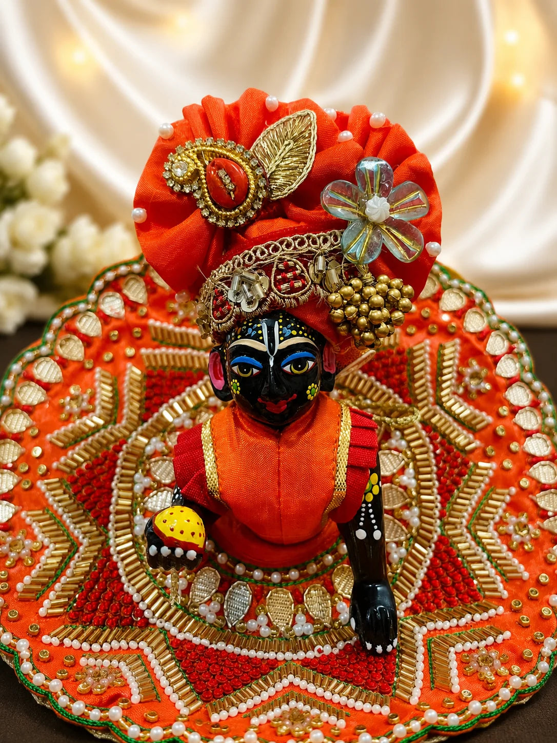 Vibrant orange festive Laddu Gopal poshak set with intricate white pearl beadwork, heavy zardozi embroidery, matching patka, and decorated turban displayed flat on white silk