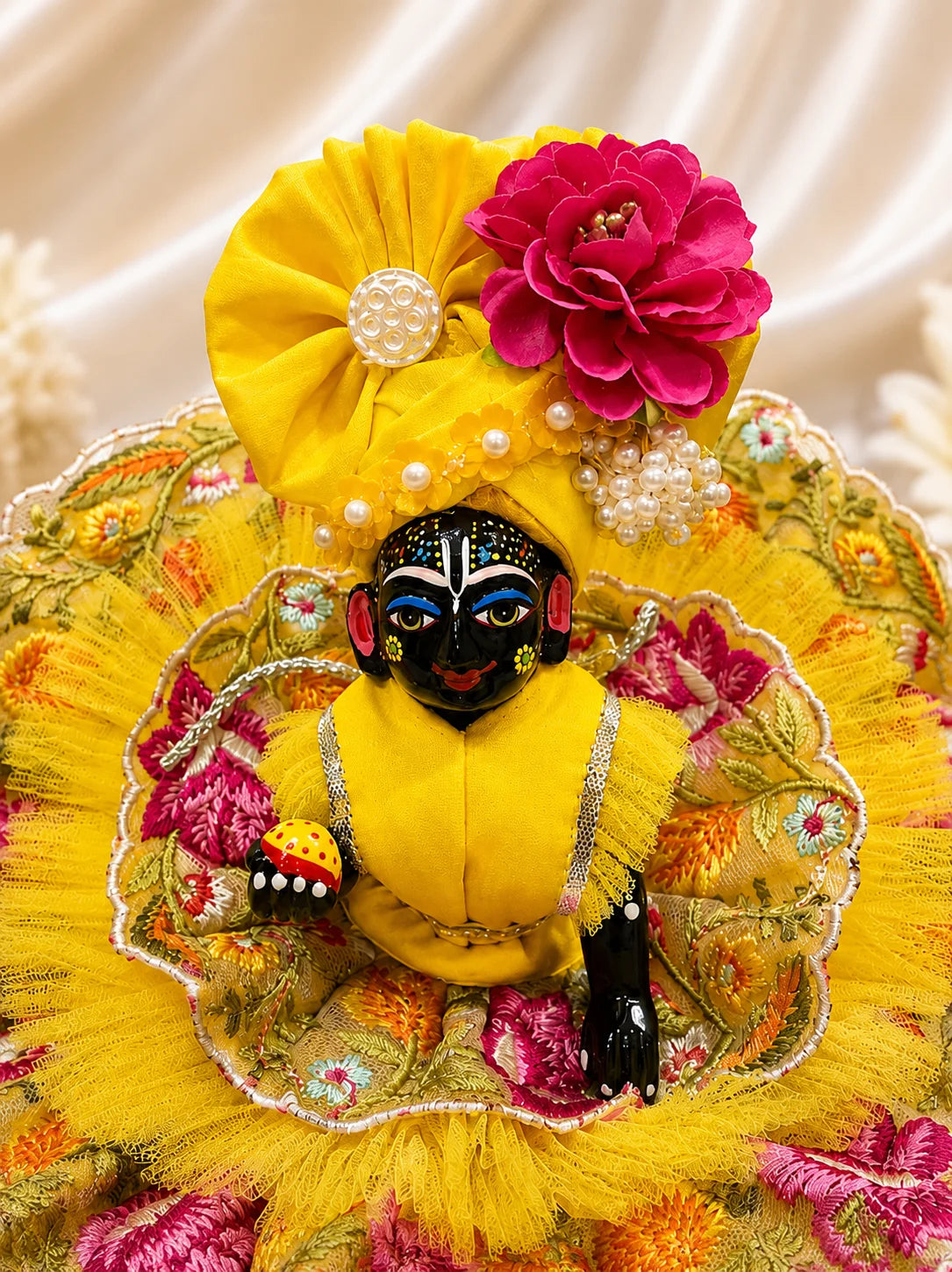 Black stone Laddu Gopal Ji idol wearing a premium yellow double-layered dress with multi-color magenta and orange floral embroidery, silver scalloped lace, and a matching yellow turban decorated with a heavy magenta fabric rose