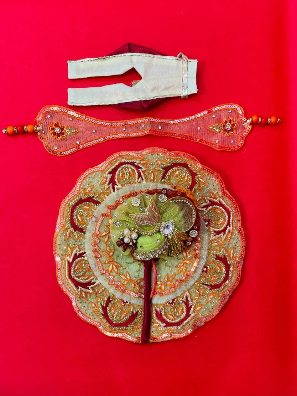 Green and orange embroidered Laddu Gopal dress set with pagdi and patka, displayed on red background and worn by idol.