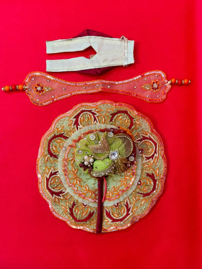 Green and orange embroidered Laddu Gopal dress set with pagdi and patka, displayed on red background and worn by idol.