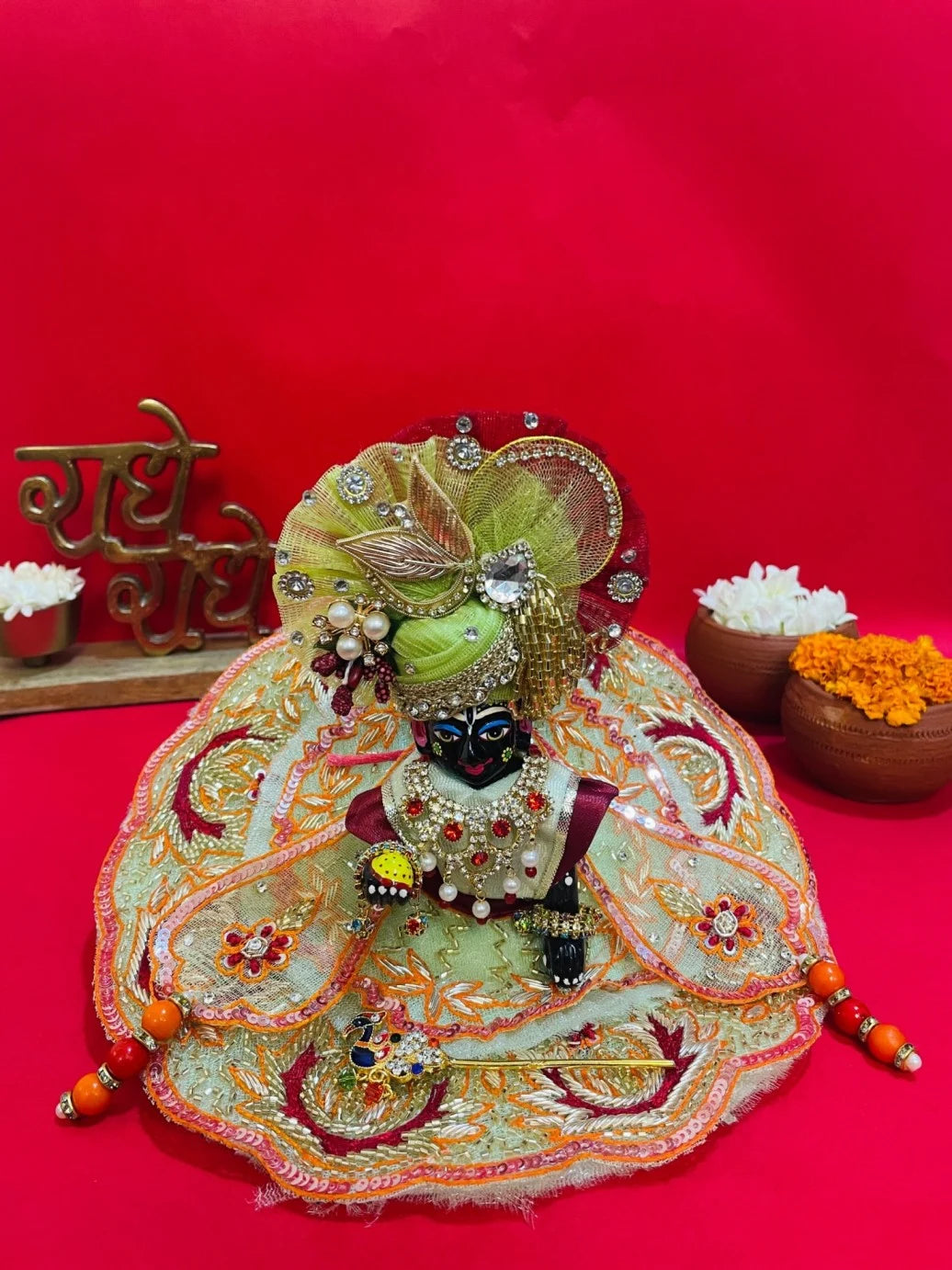 Green and orange embroidered Laddu Gopal dress set with pagdi and patka, displayed on red background and worn by idol.