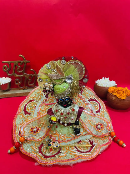 Green and orange embroidered Laddu Gopal dress set with pagdi and patka, displayed on red background and worn by idol.