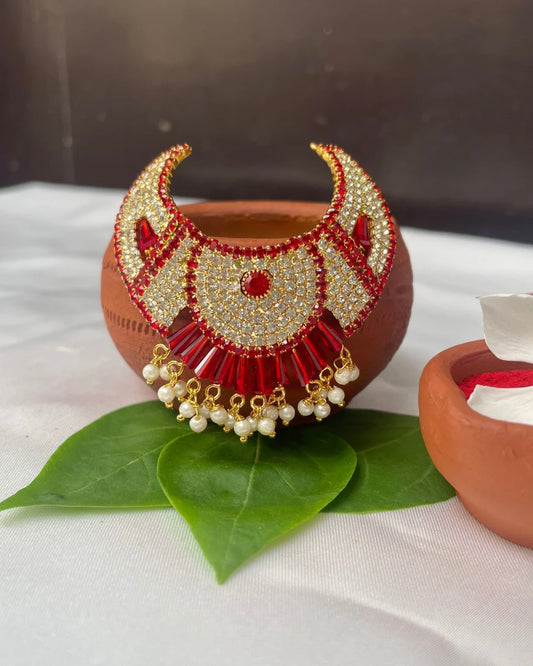 Fan-style red Laddu Gopal jewellery with white stones and pearl hangings placed on green leaves.